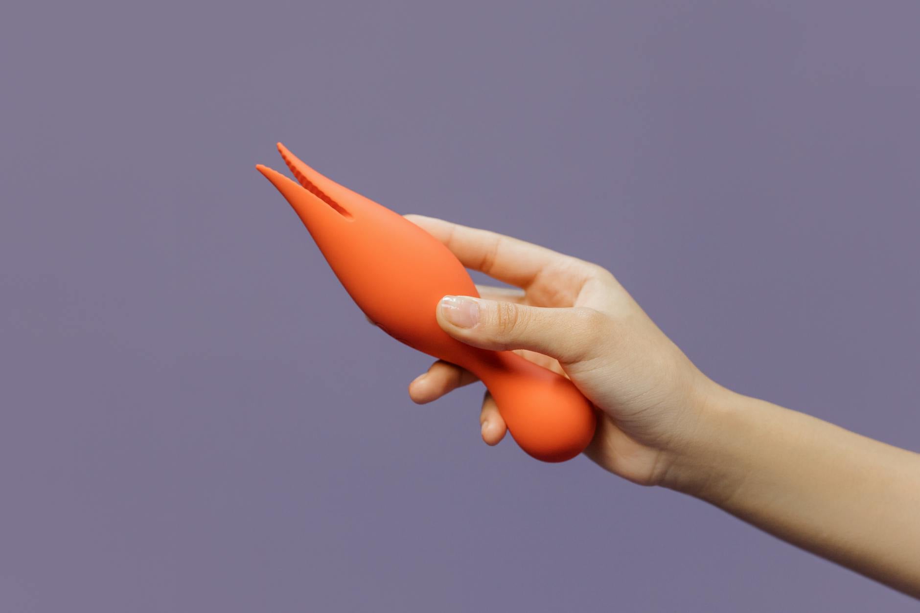 Eine Hand hält einen modernen orangen Vibrator gegen einen minimalistischen violetten Hintergrund und zeigt zeitgenössische Sinnlichkeit.