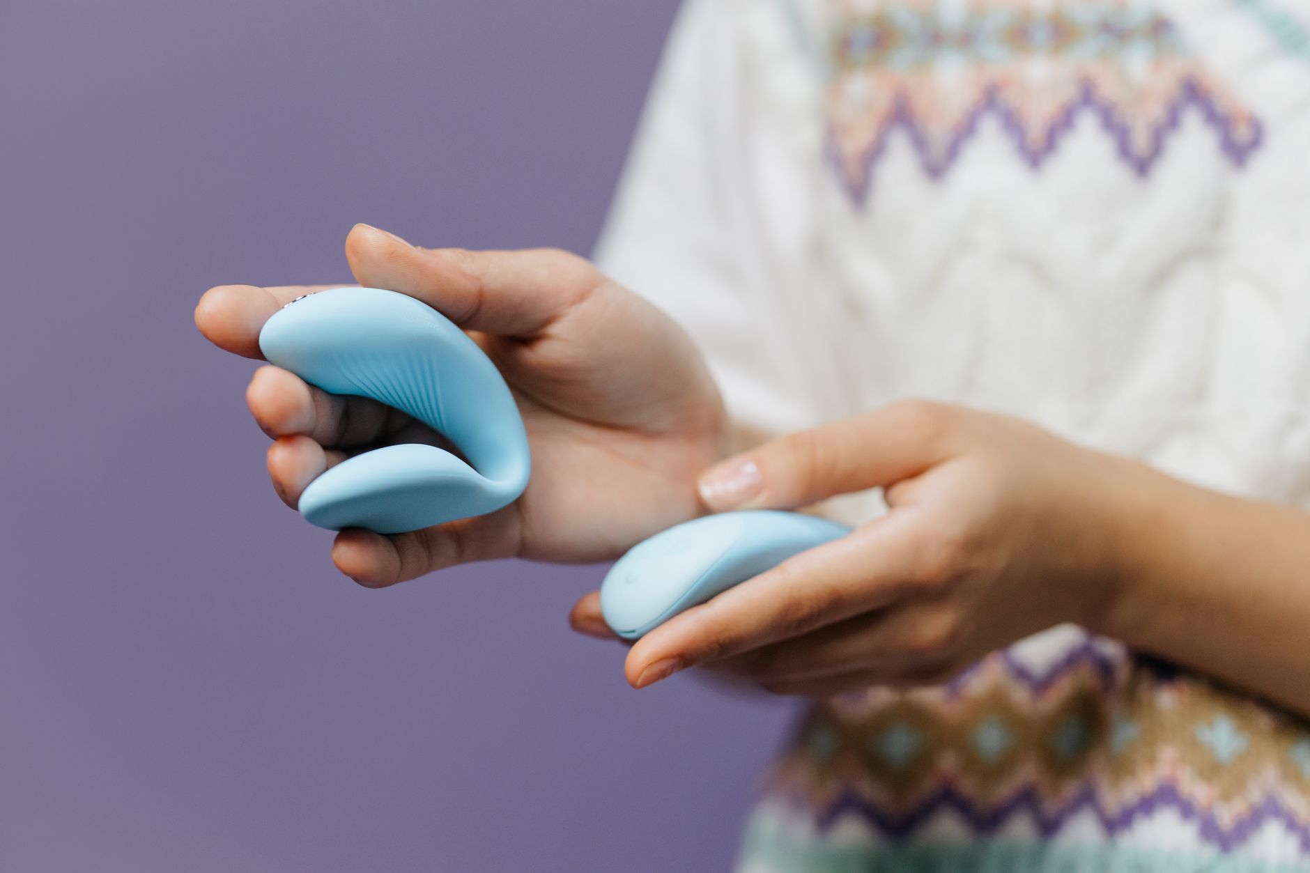 Eine Nahaufnahme zeigt Hände, die einen schlanken blauen Vibrator gegen einen violetten Hintergrund halten.
